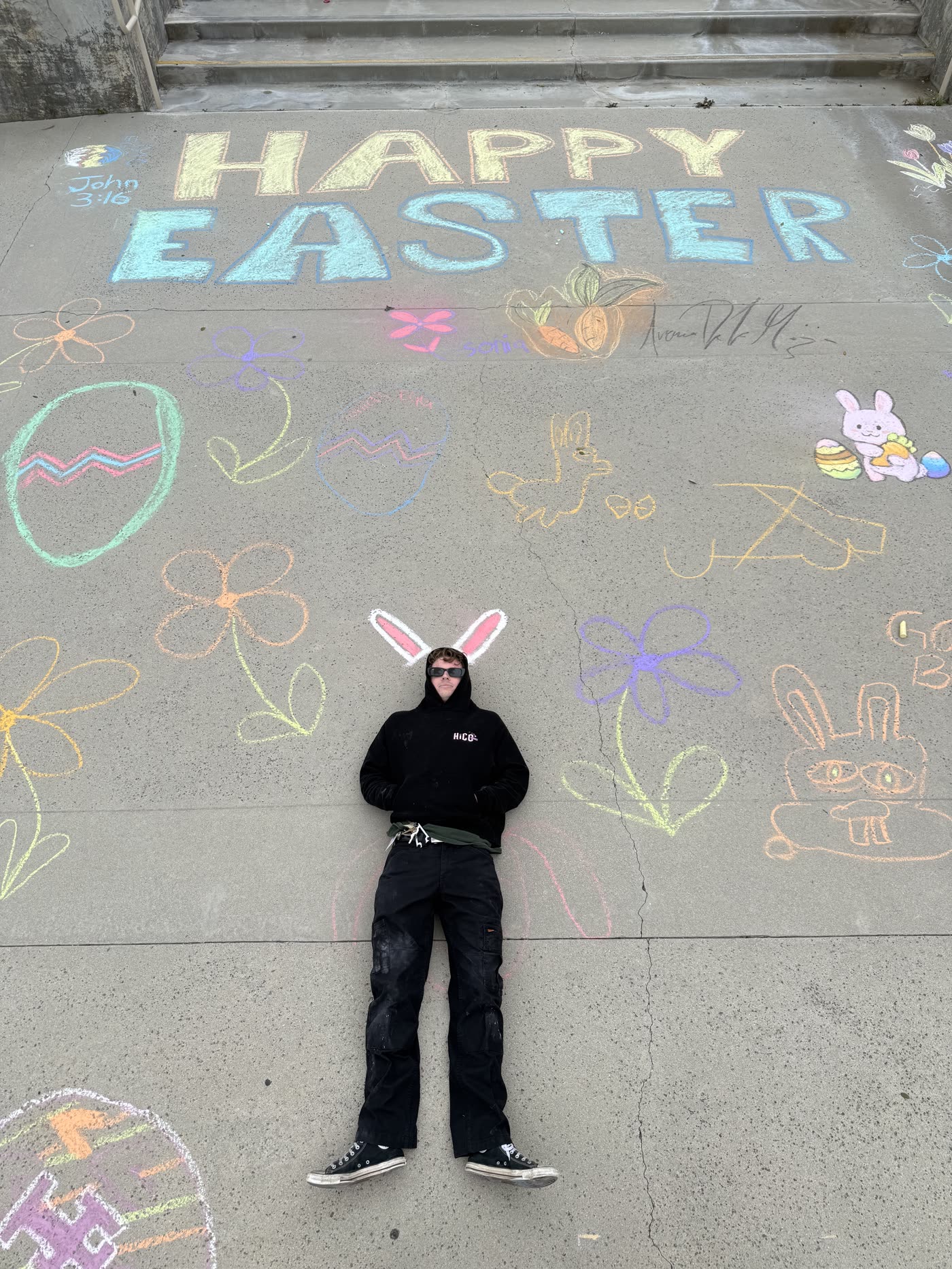 A large chalk mural reading Happy Easter with a person lying in the middle wearing rabbit ears.
